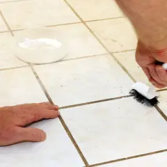 Limpiando las juntas de baldosas con un cepillo y un recipiente con polvo blanco.