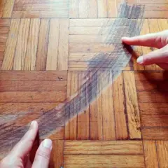 Manos señalan una mancha oscura en un suelo de parquet de madera. Se muestra cómo disimular manchas en parquet.