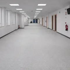 Pasillo largo y luminoso de un colegio, con ventanas a la izquierda y puertas a la derecha. El suelo del colegio es de un material gris moteado.