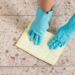 Manos con guantes azules limpiando suelos de terrazo con un paño amarillo.