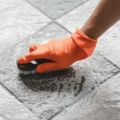 Mano con guante naranja cepillando un suelo antideslizante con espuma.