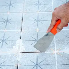 Mano con espátula naranja retirando juntas de baldosas sueltas.