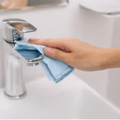 Mano limpiando el grifo cromado del lavabo con un paño azul. La limpieza del baño es esencial para un hogar saludable.