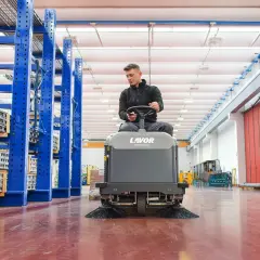 Hombre operando una maquina limpieza industrial Lavor en un almacén con estanterías azules.