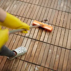 Persona con guantes amarillos y chanclas blancas usa cepillo para quitar manchas de vinagre en el suelo de madera.