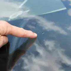Dedo señala un pequeño impacto en el parabrisas, un cristal manchado por dentro por la suciedad y el reflejo del cielo nublado.