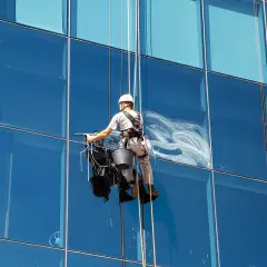 Trabajador en arnés limpiando cristales altos de un edificio moderno.