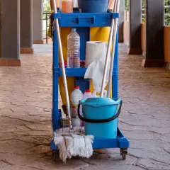 Carro de limpieza con cubeta azul, botellas y escoba sobre un suelo poroso de piedra.