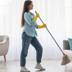 Mujer sonriente con guantes amarillos friega el suelo con una fregona. A su lado, botellas de lejía, para que sirve la lejía: para limpiar y desinfectar.