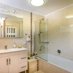 Baño con azulejos beige y detalles de mosaico. Un lavabo blanco, espejo y ducha con mampara de cristal. Ideal para aprender como poner azulejos en un baño.