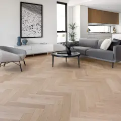 Elegante salón con suelo de madera en espiga, sofá gris, sillón y cocina moderna al fondo. La colocación espiga resalta la calidez del espacio.