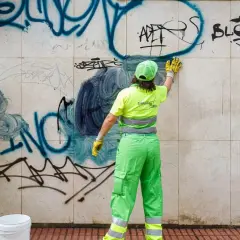 Dos trabajadores con uniformes verdes brillantes realizan la limpieza de grafitis en una pared de mármol.