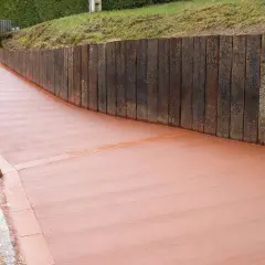 Camino de hormigón cepillado con borde de ladrillo, flanqueado por un muro de traviesas de madera.