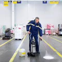 Hombre realiza limpieza almacen con pulverizador. A su lado, un bidón de líquido y una fregadora industrial.