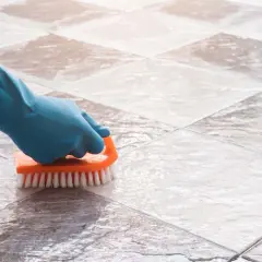 Mano con guante azul limpiando suelo de baldosas con cepillo naranja. Solución para con que fregar el suelo si no tengo fregasuelos.