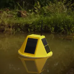 Boya amarilla con paneles solares y antena flotando en agua turbia, parte de un sistema de control de nivel de agua.