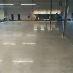 Amplio espacio industrial con suelo de **cemento pulido** impecable, reflejando las luces del techo. Se aprecian montones de tierra y maquinaria en el fondo.