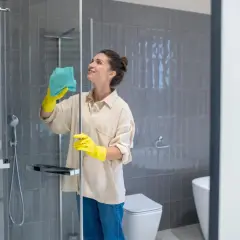 Mujer con guantes amarillos limpiando mampara ducha muy sucia con paño azul. Baño moderno con azulejos grises y sanitario blanco.
