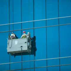 Dos trabajadores realizan limpieza en altura en una plataforma suspendida, brillando los cristales de un edificio azul.