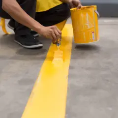 Persona pinta una línea amarilla en suelo de cemento sin pulir, usando una brocha y un cubo de pintura.