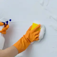 Manos con guantes naranjas rociando y limpiando paredes con espuma y esponja.