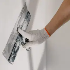 Mano enguantada aplicando material para revocar pared con llana.