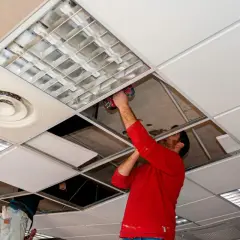 Un hombre con camisa roja instala un panel en **techos registrables**, junto a luminarias y rejillas de ventilación.