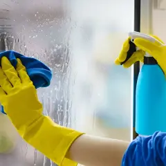 Manos con guantes amarillos limpian cristales muy sucios con un paño azul y spray.