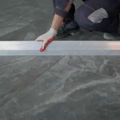 Man con guantes blancos y rojos coloca una regla metálica sobre azulejos rectificados, evitando problemas de desnivel.