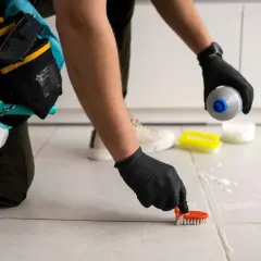 Persona con guantes negros usa un cepillo para limpiar juntas de azulejos, aplicando producto de limpieza.