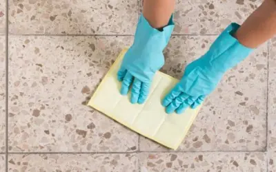 Manos con guantes azules limpiando suelos de terrazo con un paño amarillo.