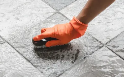 Mano con guante naranja cepillando un suelo antideslizante con espuma.