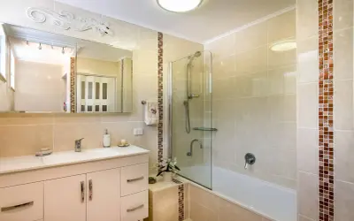 Baño con azulejos beige y detalles de mosaico. Un lavabo blanco, espejo y ducha con mampara de cristal. Ideal para aprender como poner azulejos en un baño.