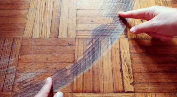 Manos señalan una mancha oscura en un suelo de parquet de madera. Se muestra cómo disimular manchas en parquet.
