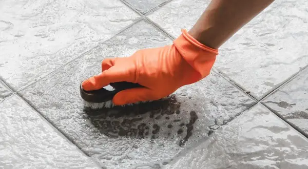 Mano con guante naranja cepillando un suelo antideslizante con espuma.