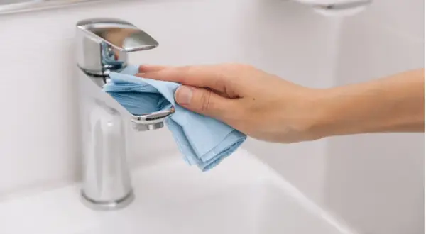 Mano limpiando el grifo cromado del lavabo con un paño azul. La limpieza del baño es esencial para un hogar saludable.