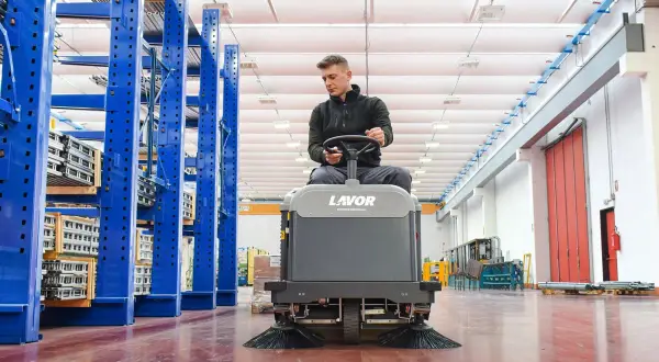 Hombre operando una maquina limpieza industrial Lavor en un almacén con estanterías azules.