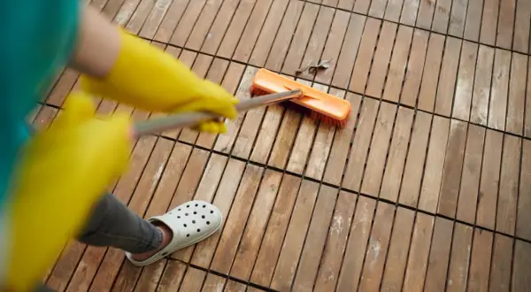 Persona con guantes amarillos y chanclas blancas usa cepillo para quitar manchas de vinagre en el suelo de madera.