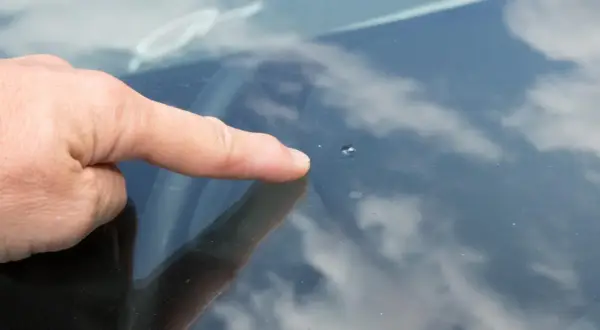 Dedo señala un pequeño impacto en el parabrisas, un cristal manchado por dentro por la suciedad y el reflejo del cielo nublado.
