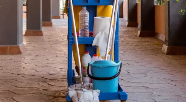 Carro de limpieza con cubeta azul, botellas y escoba sobre un suelo poroso de piedra.