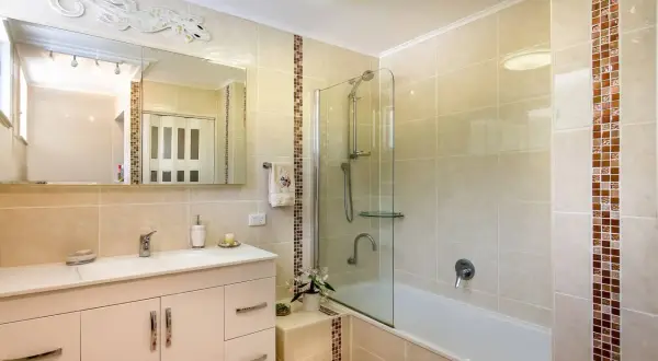 Baño con azulejos beige y detalles de mosaico. Un lavabo blanco, espejo y ducha con mampara de cristal. Ideal para aprender como poner azulejos en un baño.