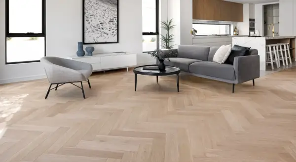 Elegante salón con suelo de madera en espiga, sofá gris, sillón y cocina moderna al fondo. La colocación espiga resalta la calidez del espacio.