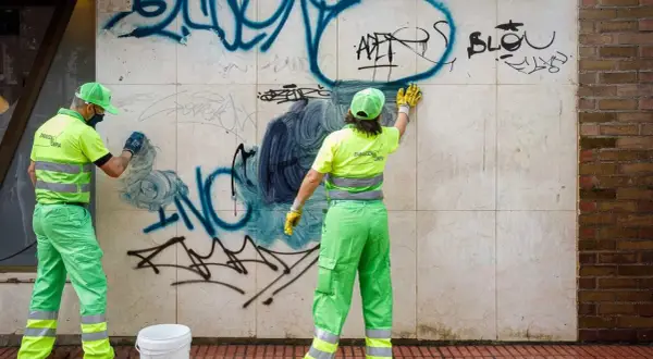 Dos trabajadores con uniformes verdes brillantes realizan la limpieza de grafitis en una pared de mármol.