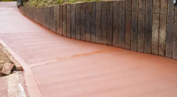 Camino de hormigón cepillado con borde de ladrillo, flanqueado por un muro de traviesas de madera.
