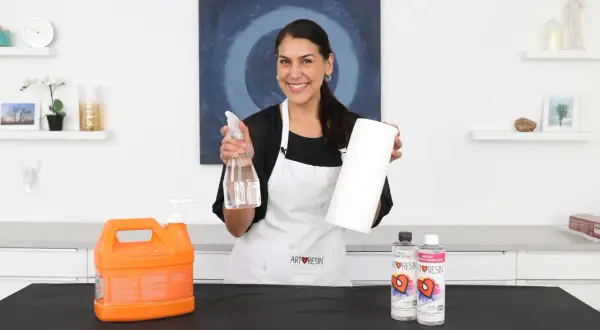Mujer sonriente muestra cómo limpiar la resina epoxi de los utensilios con spray y papel.