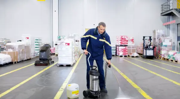 Hombre realiza limpieza almacen con pulverizador. A su lado, un bidón de líquido y una fregadora industrial.