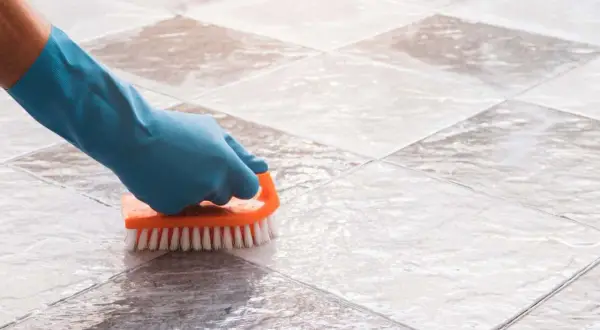 Mano con guante azul limpiando suelo de baldosas con cepillo naranja. Solución para con que fregar el suelo si no tengo fregasuelos.