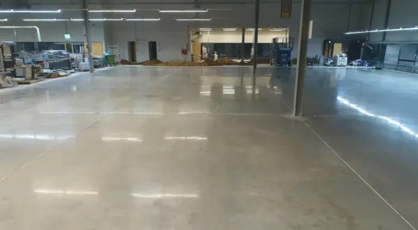 Amplio espacio industrial con suelo de **cemento pulido** impecable, reflejando las luces del techo. Se aprecian montones de tierra y maquinaria en el fondo.