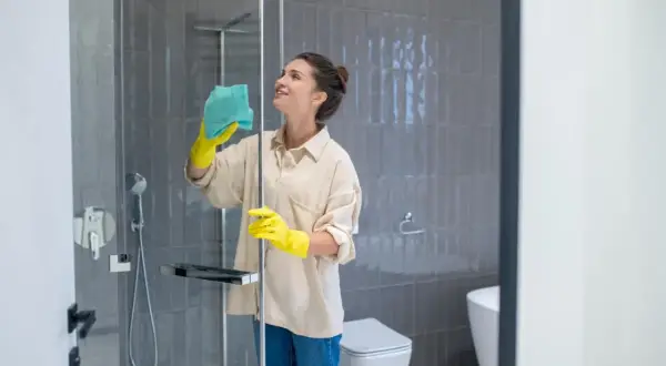 Mujer con guantes amarillos limpiando mampara ducha muy sucia con paño azul. Baño moderno con azulejos grises y sanitario blanco.