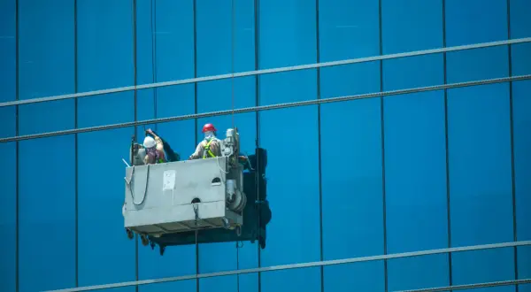 Dos trabajadores realizan limpieza en altura en una plataforma suspendida, brillando los cristales de un edificio azul.